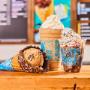 Waffle cone, shake and sundae in front of Ben & Jerry's ice cream shop menu board.