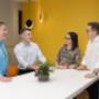 Trekk Advisory Brisbane team members having a casual stand-up meeting in a bright, colourful break-out space with a yellow feature wall and modern lighting.