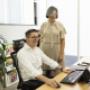 A light moment between two Trekk Advisory Brisbane team members. One’s at the desk navigating a project on screen, while the other stands nearby, sharing a laugh and offering support. Bright space, positive energy.