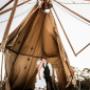 photo by Thierry Boudan Photography captures a bride and groom in a moment of joyful surprise.  Their wedding teepee has collapsed in the wind, draping them in fabric, but they laugh and embrace amidst the unexpected chaos.