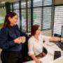 Two female professionals collaborating in Trekk Rockhampton office. One is seated and pointing at a computer screen, while the other stands beside her with a coffee mug, both engaged in discussion.