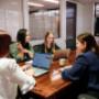 Four team members of Trekk Rockhampton office in a collaborative meeting around a conference table. They're engaged in conversation with laptops open and coffee cups on hand. A smart TV screen is visible in the background.
