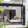 Covered outdoor terrace with louvered windows, built-in kitchenette, and gray sectional overlooking trees.
