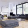 Spacious living room with long dark sectional, fireplace, floor-to-ceiling windows, and light wood floors.
