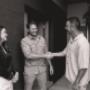 Black-and-white photo of two men shaking hands at a doorway while a woman smiles beside them.